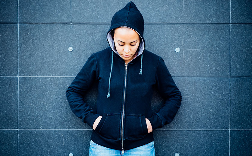 Young black girl in hoodie and jeans leans against wall, eyes down