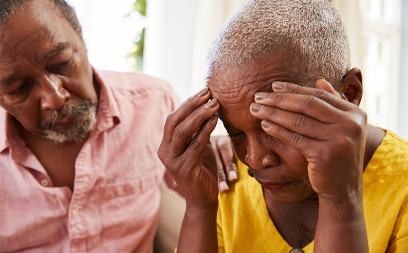 An African American couple sits on a couch. The woman looks upset, head in hands as he comforts her.