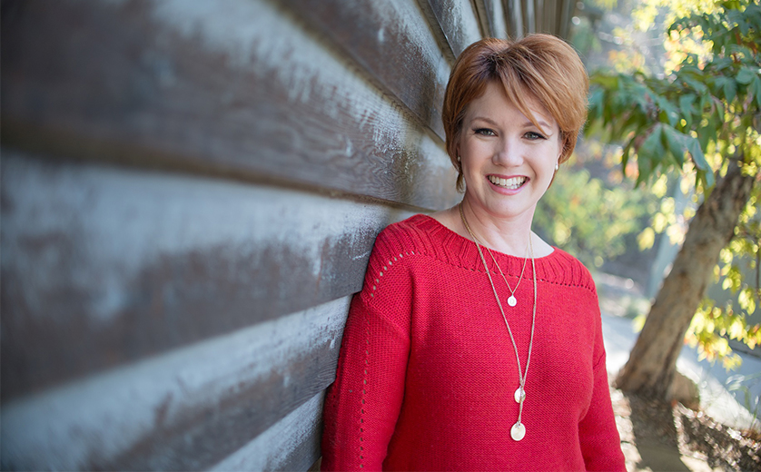 Smiling woman in a red sweater leaning against a wooden wall outdoors on a sunny day