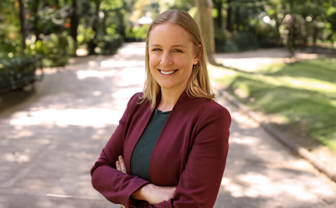 Professional woman with arms crossed, smiling on a sunlit park path in a burgundy blazer.