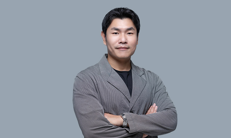 Professional portrait of a man with arms crossed, wearing a gray blazer and black shirt, standing against a light blue-gray background.