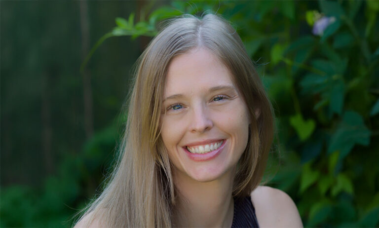 Portrait of a smiling woman with long blonde hair, standing outdoors with greenery in the background.