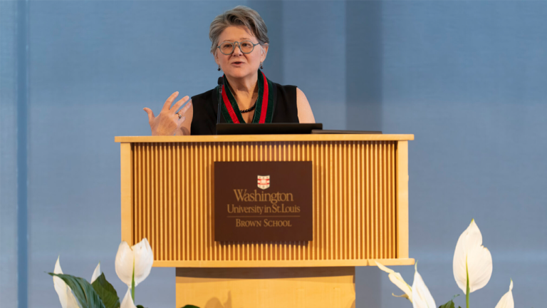 Woman in glasses speaks at a wooden podium with a Washington University in St. Louis Brown School plaque, blue backdrop behind her.