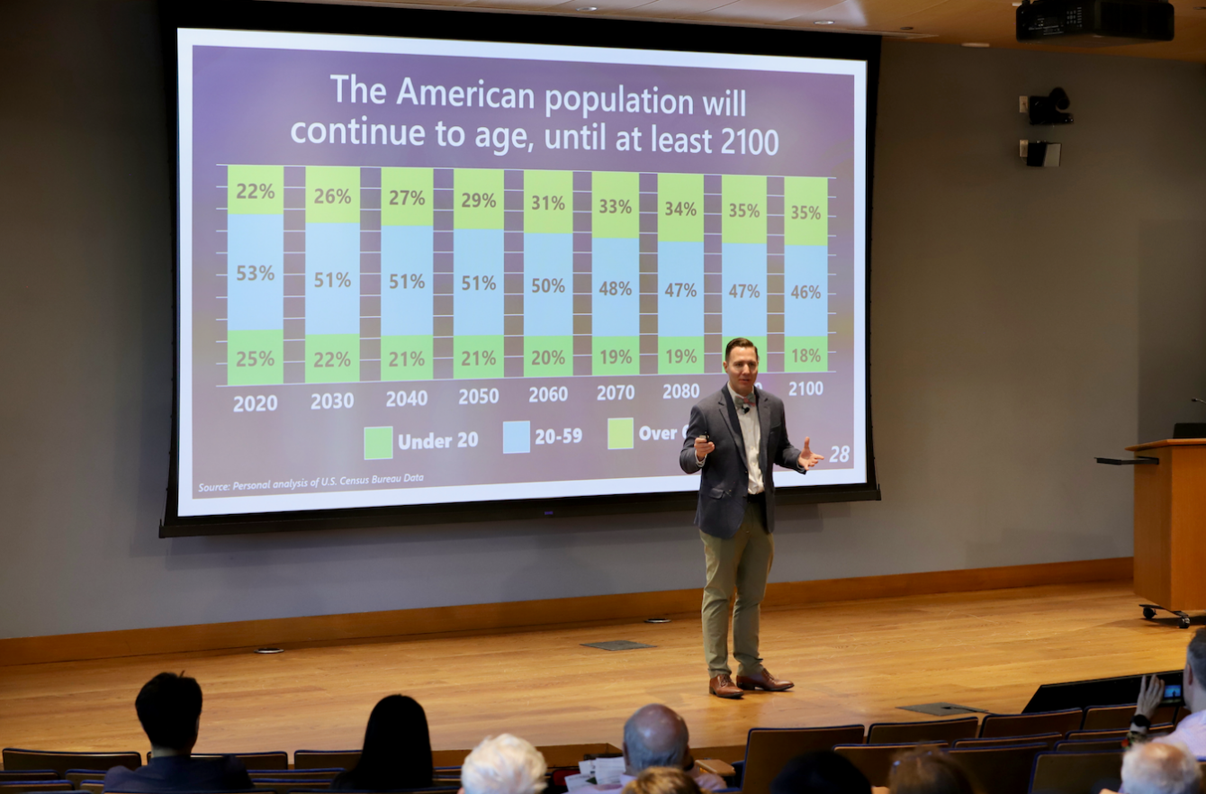 Presenter in a lecture hall giving a talk with a projected slide showing a stacked bar chart of US age distribution from 2020 to 2100 (Under 20, 20–59, Over 60).