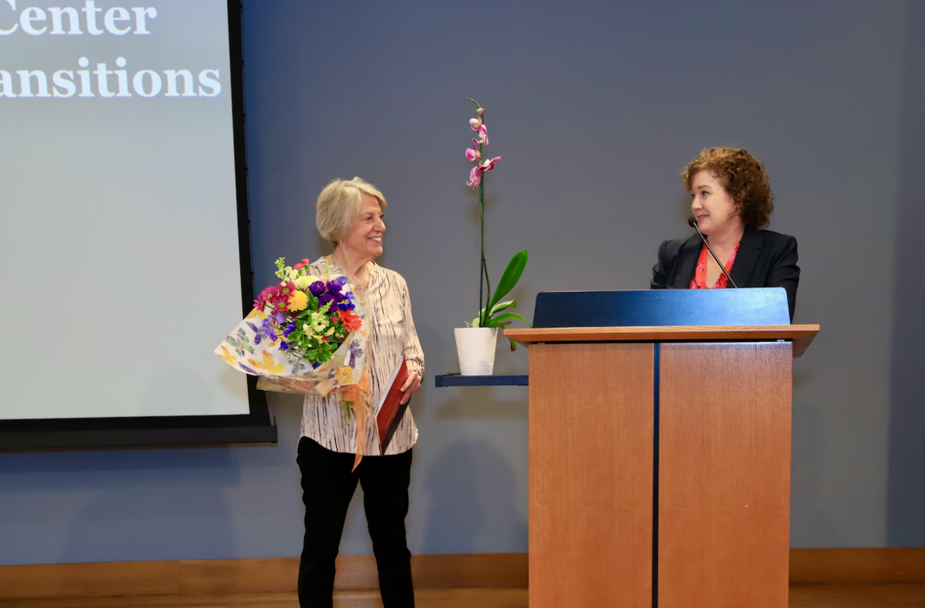 Smiling woman with short gray hair receiving a colorful bouquet and a plaque from a speaker at a podium on stage.