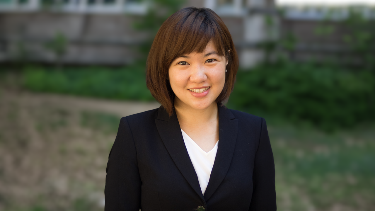 Portrait of a woman in a black blazer smiling outdoors with greenery in the background.