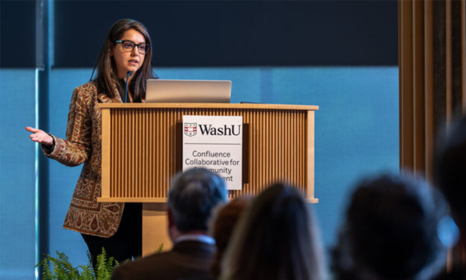 Woman with glasses speaks at a wooden podium with a WashU sign, addressing an audience in a conference room.