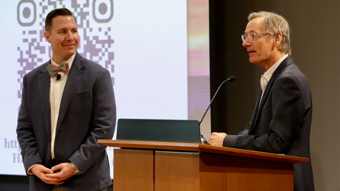 Two men in a conference room: one in a blue blazer with a bow tie stands beside a podium while the other speaks at the mic, QR code screen behind them.
