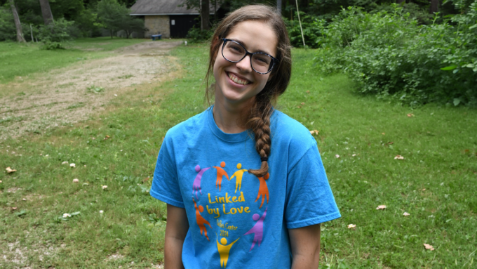 Smiling girl with glasses and a braided ponytail outdoors in a green yard, wearing a blue T-shirt that says 'Linked by Love'.