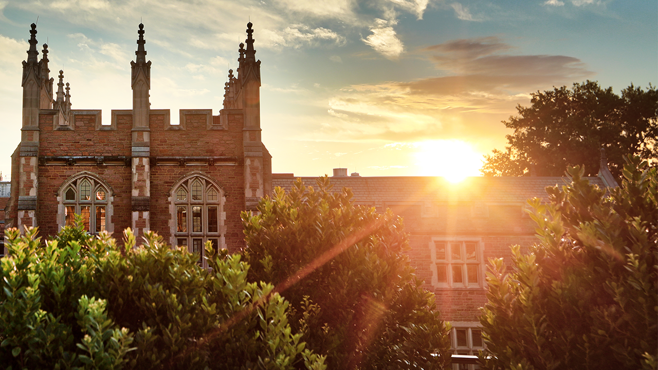 Sunset over a red brick gothic-style building with pointed spires, seen behind green shrubs in the foreground.