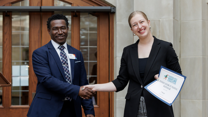 Two professionals shake hands outside a building; one holds an award certificate and smiles.