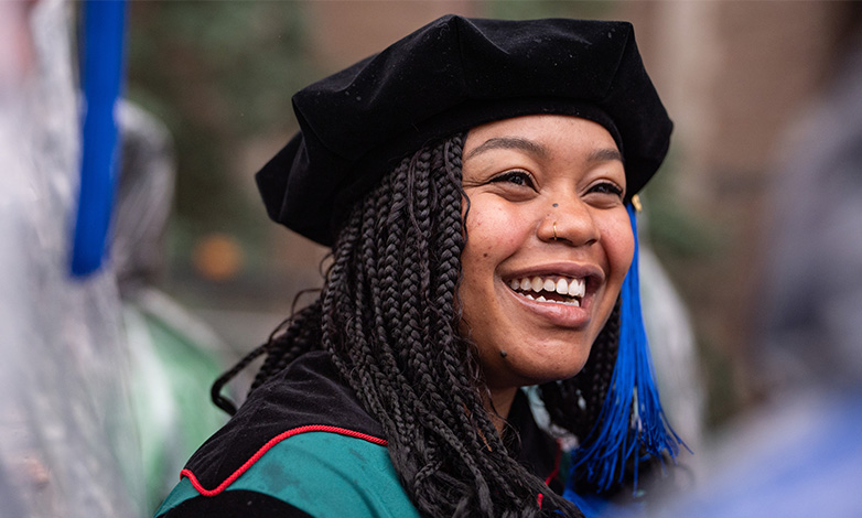 Smiling graduate wearing a black cap with a blue tassel and a teal-accented gown during a ceremony