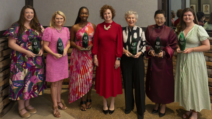 Eight women stand side by side, each holding a glass award at an awards ceremony, wearing colorful dresses and formal attire.