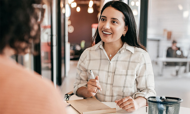 Woman with dark hair in a white plaid shirt smiles while taking notes at a table in a bright cafe.