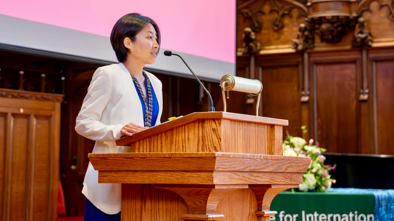 Woman in a white blazer delivers a speech at a wooden podium with a microphone in a formal venue.