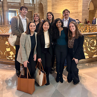 Group of eight young adults in business attire pose for a photo inside a grand historic building with a decorative gold railing on a marble floor.