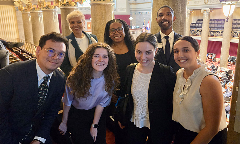 Six diverse young professionals pose for a group photo in a grand legislative chamber with marble columns and balconies, smiling at the camera.