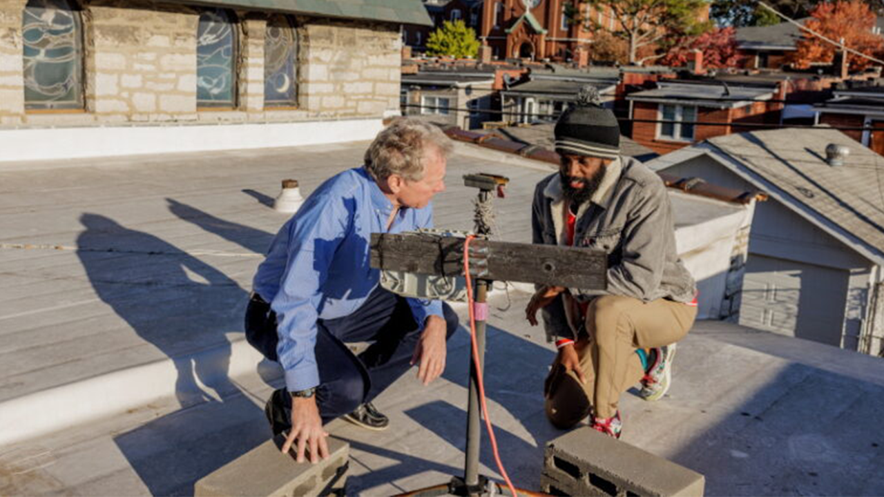 Two construction workers crouch on a residential rooftop, inspecting a makeshift support rig with a wooden beam and metal post.