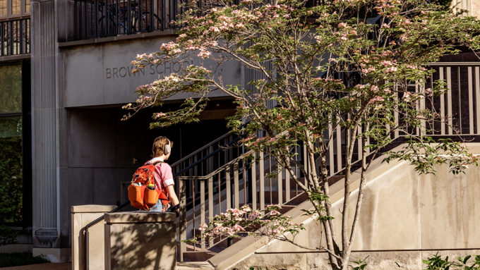 Student wearing headphones and an orange backpack walks up concrete stairs toward a building with a sign that reads 'Brown School' amid flowering trees.