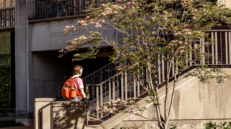 Student wearing headphones and an orange backpack walks up concrete stairs toward a building with a sign that reads 'Brown School' amid flowering trees.