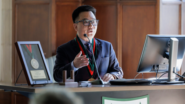 Man in a dark suit with a medal and glasses speaks into a microphone at a desk, with a framed award and computer monitor nearby.