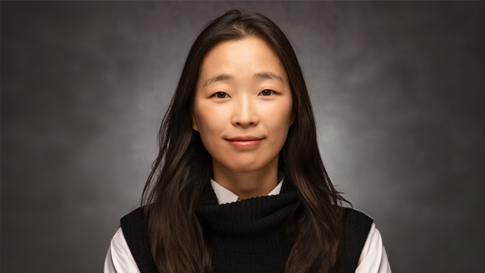 Portrait of an East Asian woman with long dark hair, wearing a black knit turtleneck over a white shirt, smiling softly against a gray backdrop.