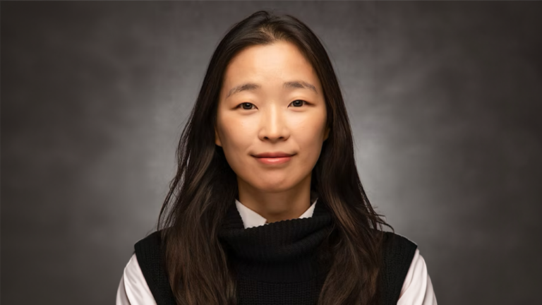 Portrait of an East Asian woman with long dark hair, wearing a black knit turtleneck over a white shirt, smiling softly against a gray backdrop.