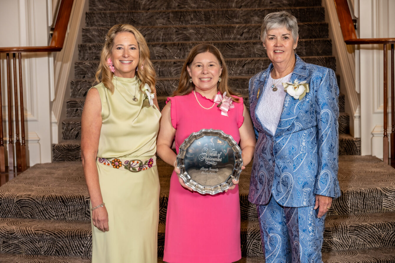 Three women in pastel dresses stand on a carpeted staircase, with the center woman in pink holding a silver award plate.