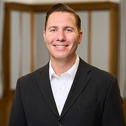 Professional headshot: smiling man in a dark suit and white shirt, indoors with a blurred wooden background