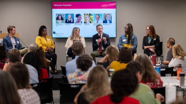 Panel discussion with six panelists seated on stage in a conference room, audience watching a presentation on a large screen behind them.