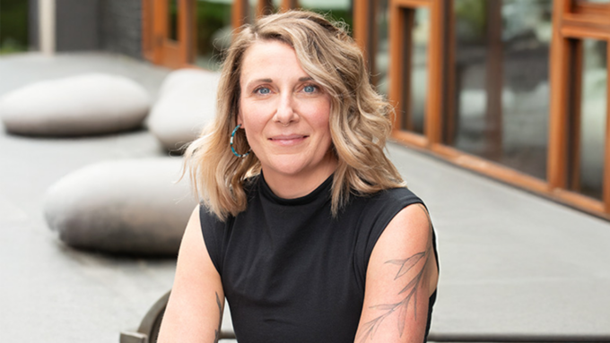 Portrait of a woman with blonde, shoulder-length hair wearing a black sleeveless top, seated outdoors with large stone features and wood-framed glass behind her, showing a relaxed smile.