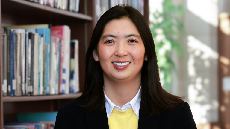 Professional headshot of a smiling woman with dark hair in a library background, wearing a blazer over a yellow sweater
