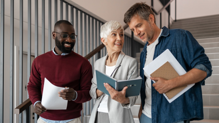 Three coworkers walking in a bright stairwell, smiling while reviewing papers together on a sunny day in the office.