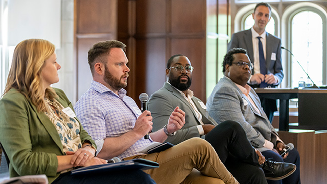 Panel discussion with four speakers seated in a row, one man holds a microphone and speaks as a moderator stands at a podium in the background.