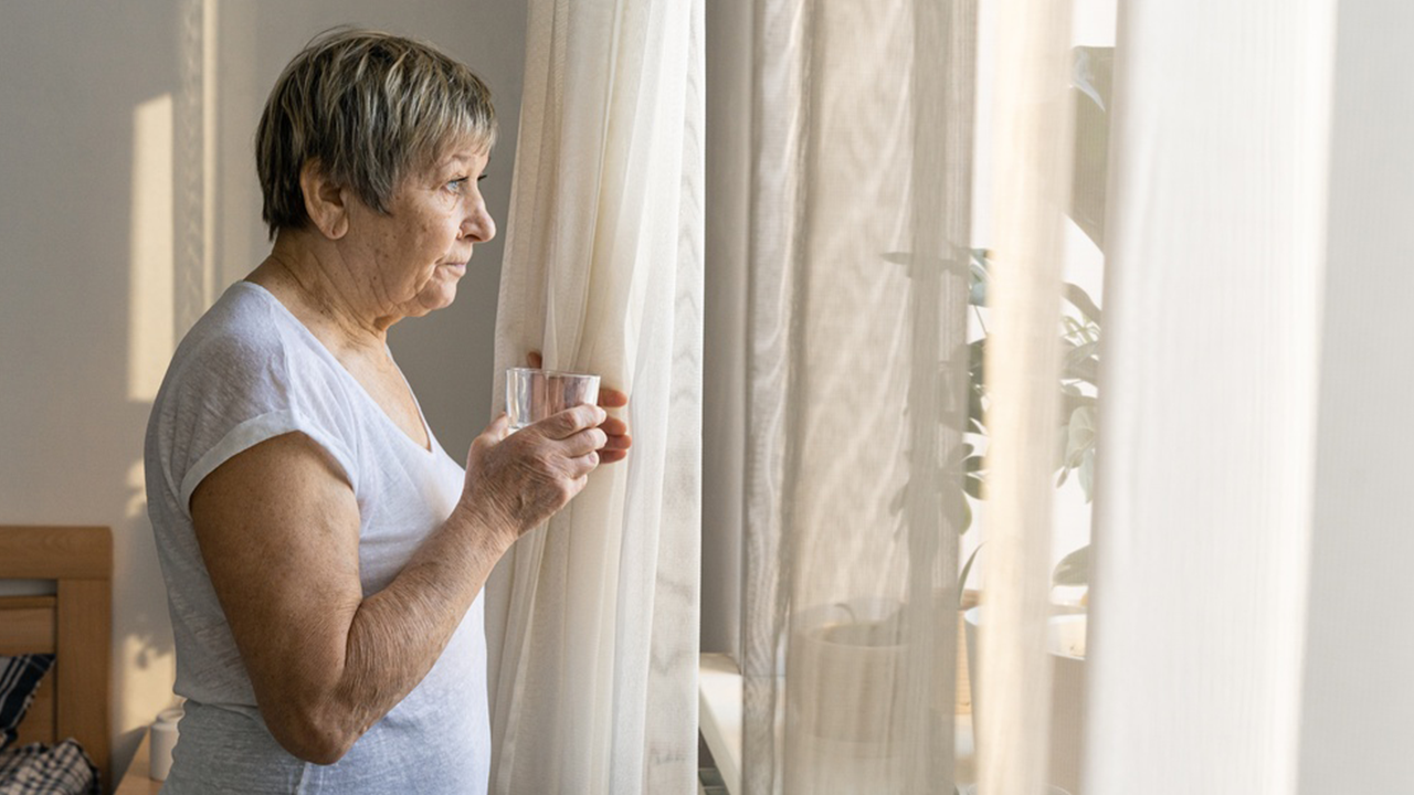 Older woman standing by a sunlit window, holding a glass of water and looking outside.