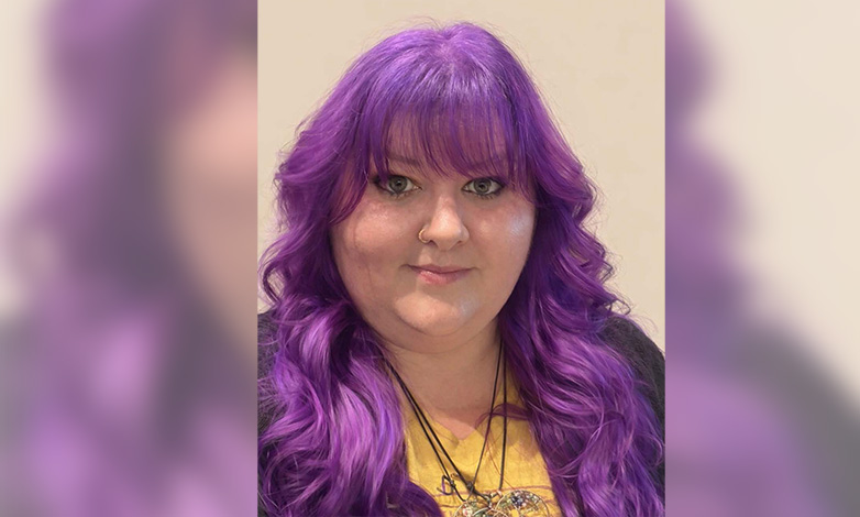 Portrait of a woman with long wavy purple hair and bangs, wearing a yellow top and a necklace, looking at the camera.