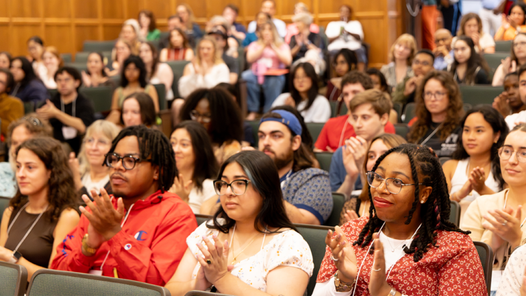 Diverse audience in a wood-paneled auditorium clapping while watching a presentation.