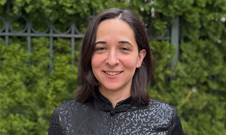 Smiling woman with shoulder-length dark hair in a dark patterned jacket, outdoors with greenery behind.