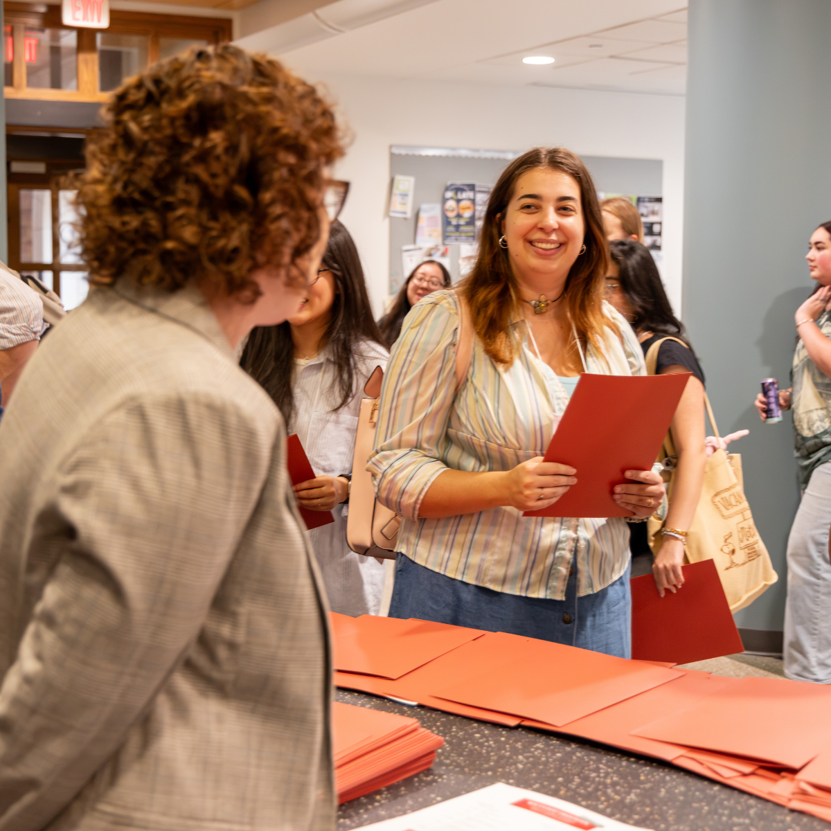 A smiling woman in a striped shirt holds a red folder at a table with more red folders, engaging with others in a event or registration setting.