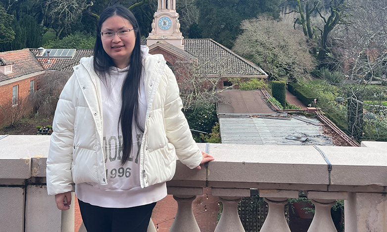 Woman with long dark hair and glasses leaning on a balcony railing, wearing a white puffer jacket, with a clock tower in the background.