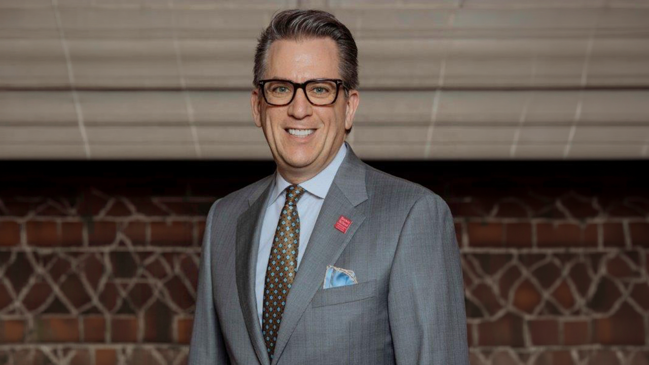 Smiling man in a gray suit and glasses, blue shirt, patterned tie, standing in front of a brick wall. While wearing a pocket square.