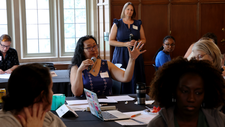 Woman with glasses speaks into a handheld microphone at a table during a workshop, raising her left hand to emphasize a point as others listen nearby.