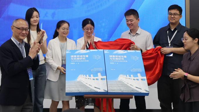 A diverse group of eight people unveiling two large blue books on a stage, with a red cloth pulled aside, smiling and clapping as the books are revealed.