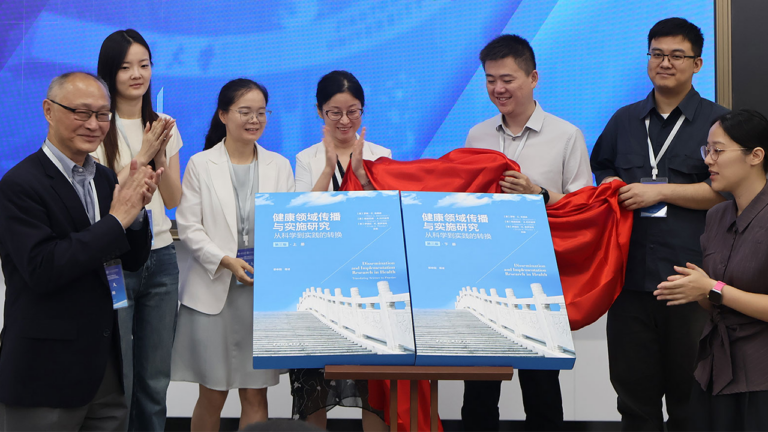 A diverse group of eight people unveiling two large blue books on a stage, with a red cloth pulled aside, smiling and clapping as the books are revealed.