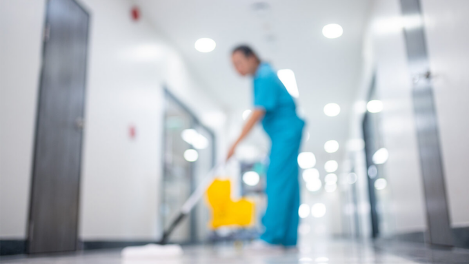 A janitor in blue scrubs mops a bright hallway, with a yellow mop bucket in the background.