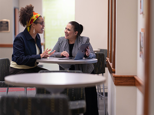 Two women sit at a round table in a bright hallway, smiling and talking; one gestures with her hands while the other holds a tablet on the table.