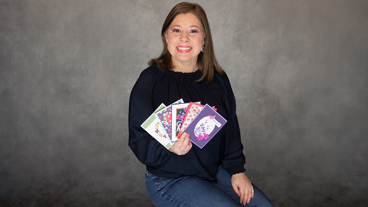 Smiling woman in a dark blouse sits against a gray backdrop, holding a fan of colorful greeting cards.