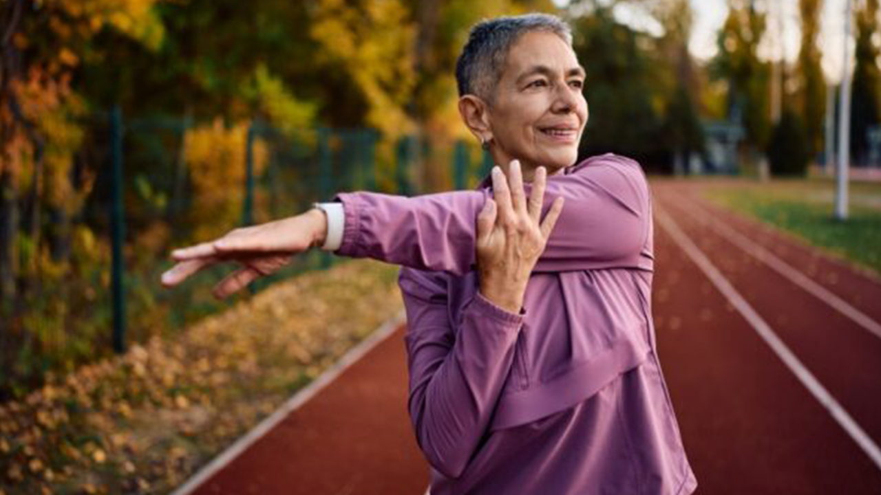 Senior woman in a pink jacket stretches her arm across her chest on an outdoor running track in autumn colors, preparing for exercise.