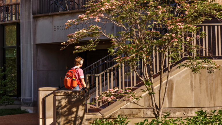 Person with orange backpack and headphones walking up stairs beside a building labeled Brown School, shaded by a flowering tree.