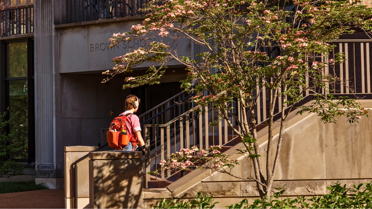 Person with orange backpack and headphones walking up stairs beside a building labeled Brown School, shaded by a flowering tree.